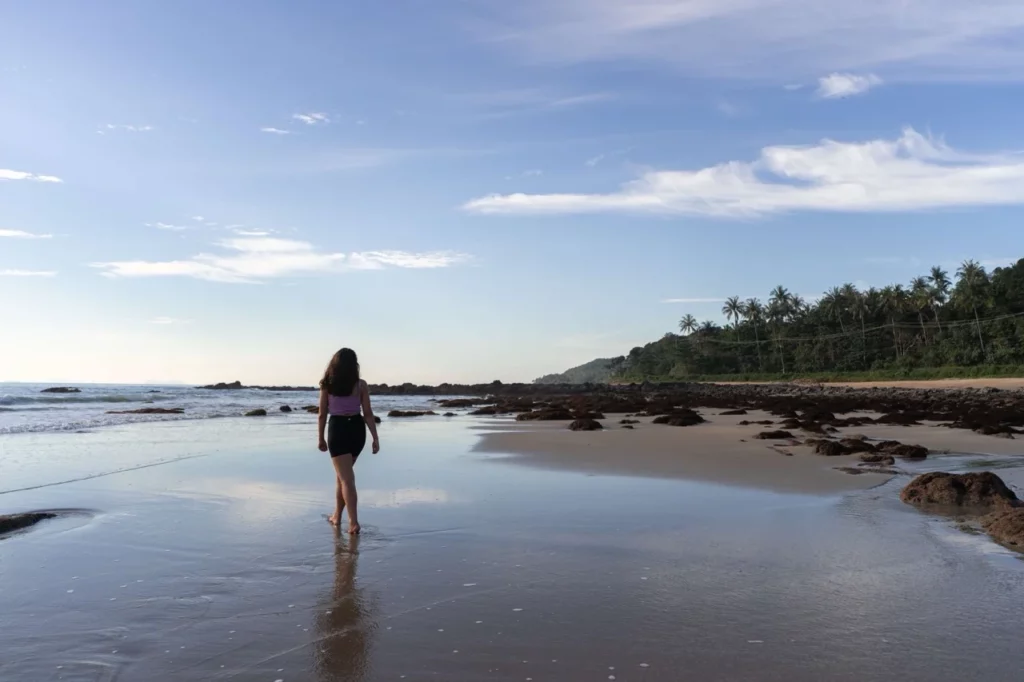 Clara spaziert an einem ruhigen Strand entlang und plant ihren Urlaub 2025 unter azurblauem Himmel und tanzenden Palmen.