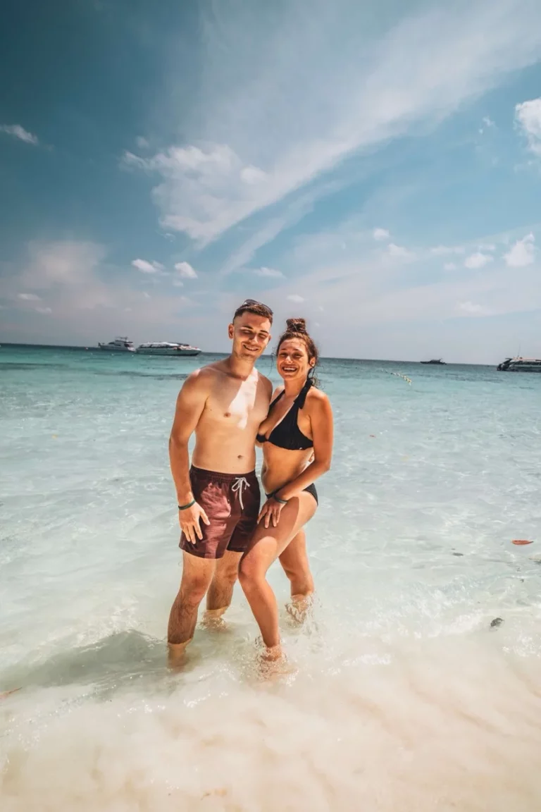 Clara und Jonathan genießen die Sonne an einem Sandstrand mit klarem Wasser und Booten und träumen vom Sommerurlaub 2025.