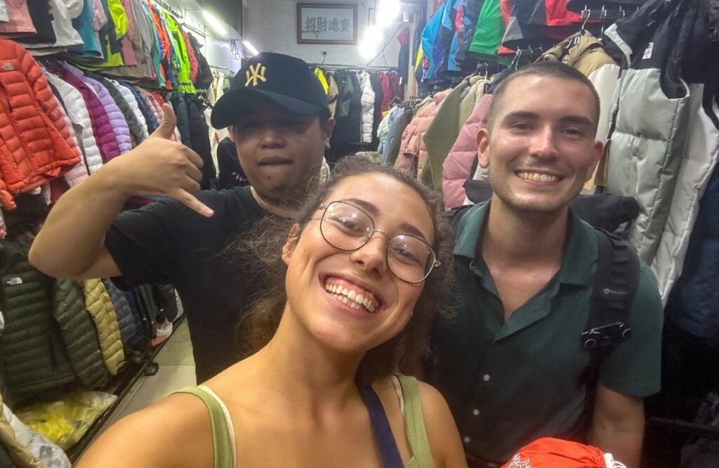 Clara und Jonathan lächeln mit einem Local auf einem Marktplatz in Ho-Chi-Minh und kaufen Ausrüstung für Vietnam Reise ein.