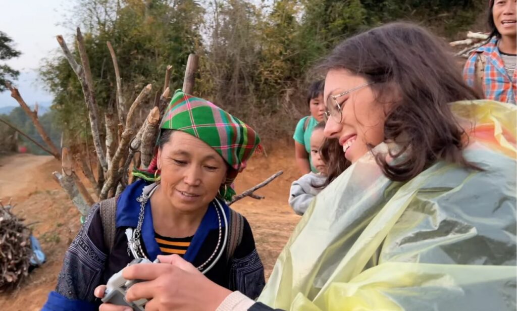 Clara steht draußen in Vietnam mit einer einheimischen Frau, die Holz trägt, während im Hintergrund ein Kind spielt.