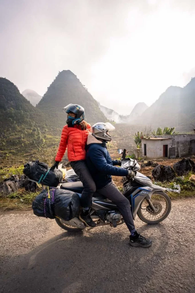 Clara und Jonathan fahren mit ihrem Gepäck unter bewölktem Himmel auf einem Motorrad durch die beeindruckenden Berge Vietnams.