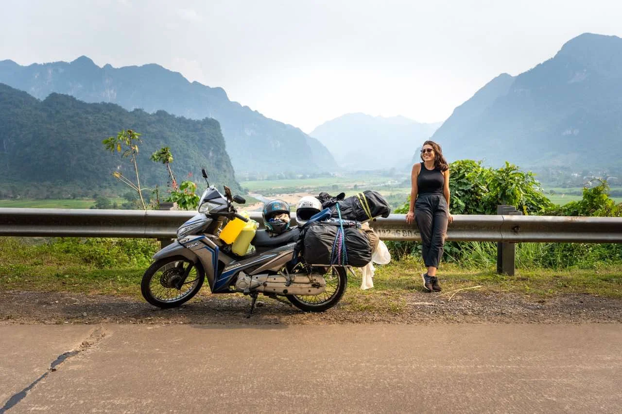 Clara steht neben einem mit Gepäck beladenen Motorrad auf Vietnams malerischer Phong Nha Nationalpark mit beeindruckenden Bergen im Hintergrund.