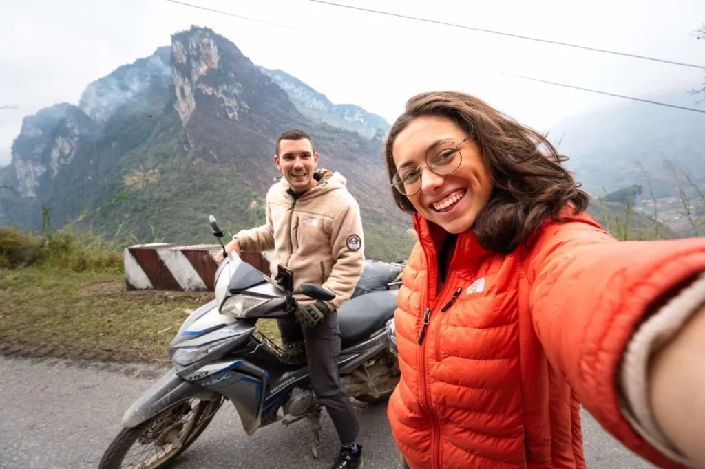 Clara und Jonathan lächeln auf der Vietnam Route, im Hintergrund sind  neblige Bergpanoramen, während sie auf dem  Motorrad sitzen.