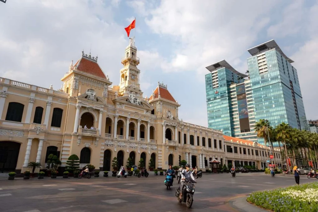 Der koloniale Uhrenturm ragt hoch in die Höhe, eingerahmt von eleganten Wolkenkratzern, an denen Motorräder vorbeirasen und die vietnamesische Flagge weht.