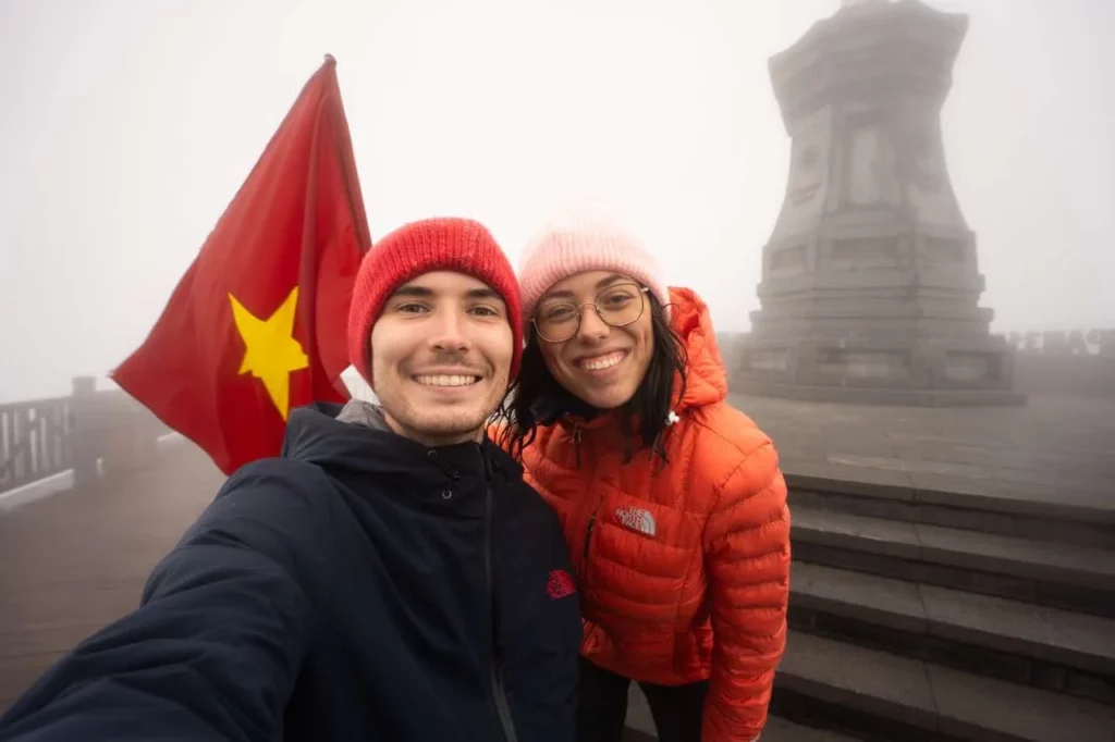 Clara und Jonathan mit der vietnamesischen Flagge im Hintergrund inmitten einer nebligen Landschaft auf dem Fansipan Berg.