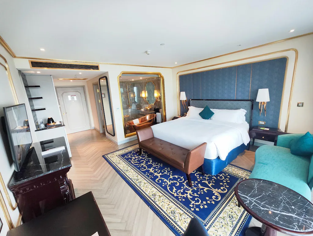 Das geräumige Hotelzimmer in Vietnam verfügt über ein bequemes Kingsize-Bett, eine elegante blaue Akzentwand und ein schickes Bad mit Glasverkleidung.