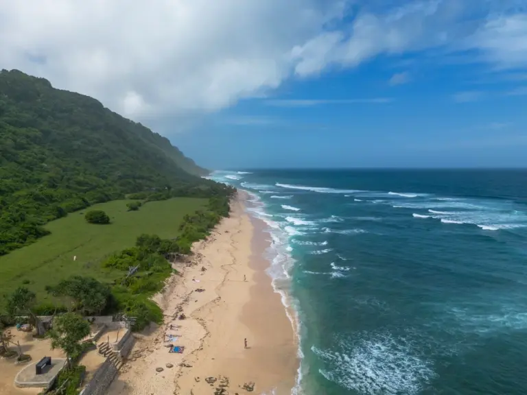 Drohnenaufnahme von einem Strand in Uluwatu mit Klippen im Hintergrund, die Wellen brechen entlang der Küste.