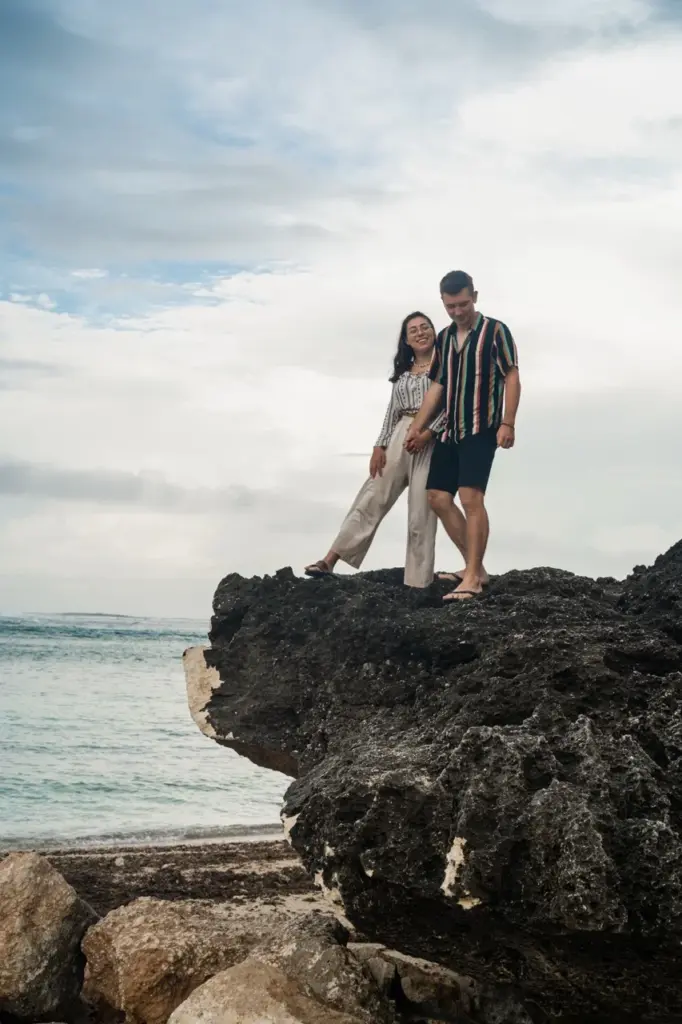Clara und Jonathan auf einem großen Felsen am Strand und hören das Rauschen der Meereswellen auf einer Bali Backpacking Reise.
