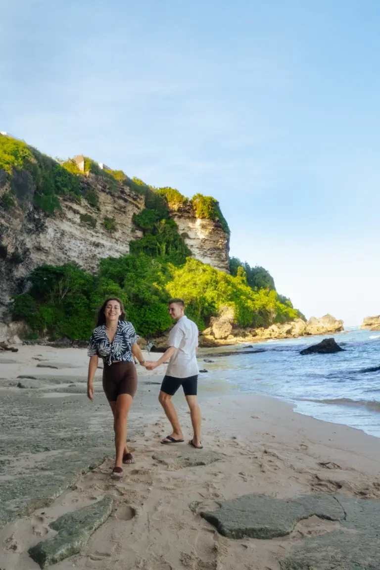 Clara und Jonathan schlendern Hand in Hand an einem sonnigen Strand auf Bali entlang, umgeben von atemberaubenden Felsklippen.