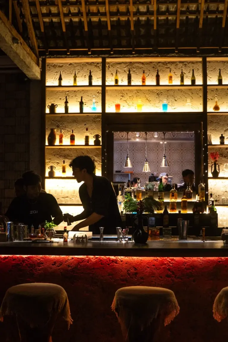 Entdecken Sie Balis pulsierendes Nachtleben, während ein Barkeeper inmitten leuchtender, flaschenbeleuchteter Regale Cocktails mixt.