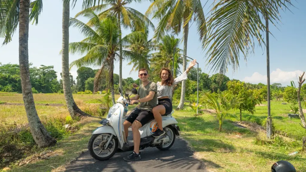 Clara und Jonathan fahren mit einem Motorroller durch Balis üppig grüne Felder, mit hohen Palmen und strahlend blauem Himmel im Hintergrund und sammeln die besten Bali Reisetipps.