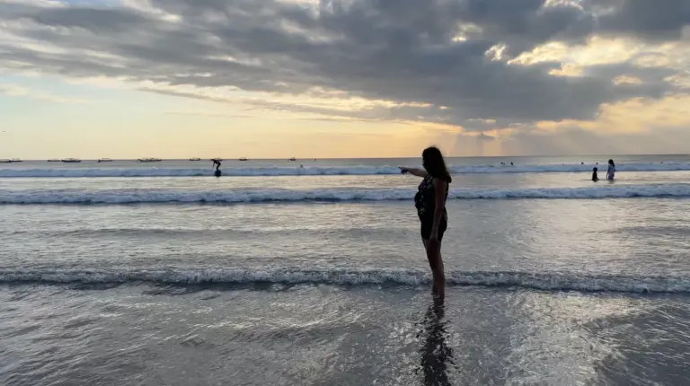 Clara bewundert vom seichten Wasser aus den leuchtenden Sonnenuntergang auf Bali, umrahmt von dramatischen Wolken und dem ruhigen Ozean.