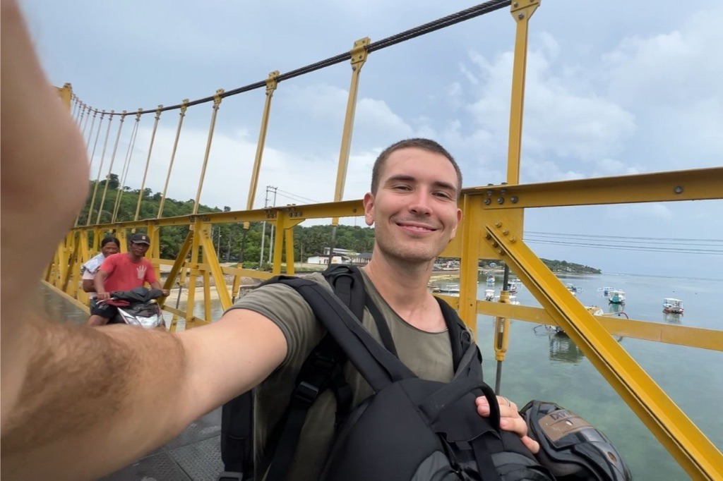 Jonathan macht ein Selfie auf Balis berühmter Gelber Brücke, während Motorräder vorbeirauschen und man das Meer im Hintergrund sieht.