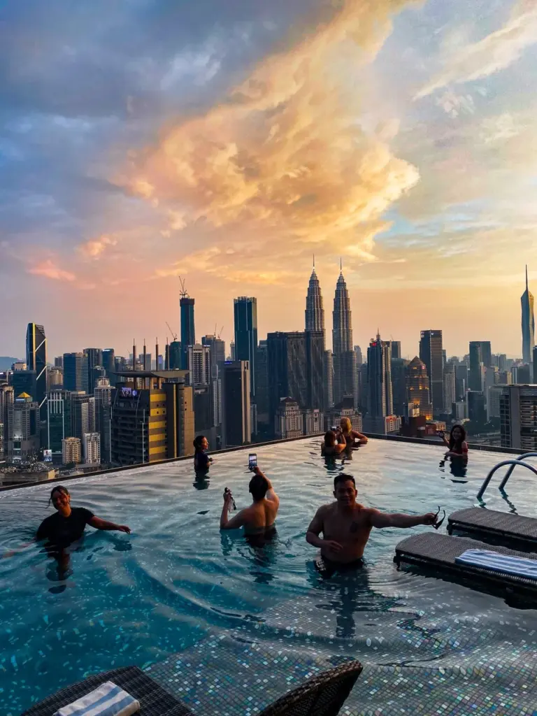 Clara und Jonathan entspannen in einem Pool auf dem Dach mit Blick auf die Skyline von Kuala Lumpur bei Sonnenuntergang.