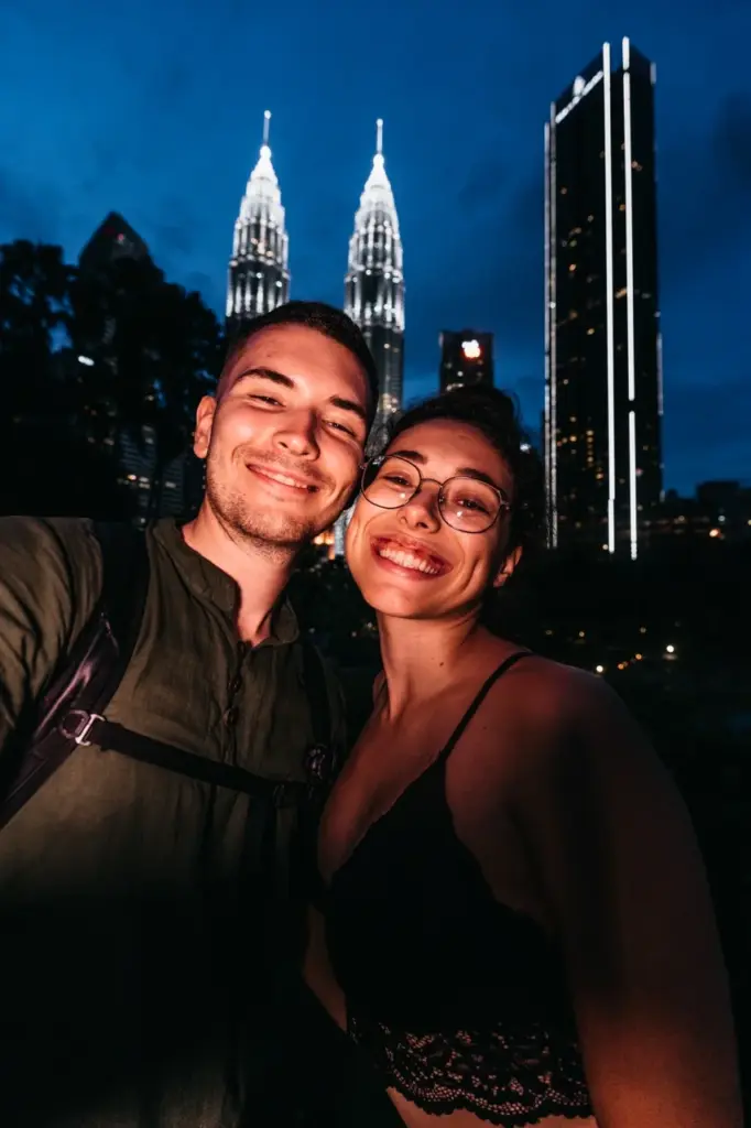 Clara und Jonathan lächeln strahlend, während hinter ihnen die schillernden Türme Kuala Lumpurs leuchten. [Stichwort: Reisen].