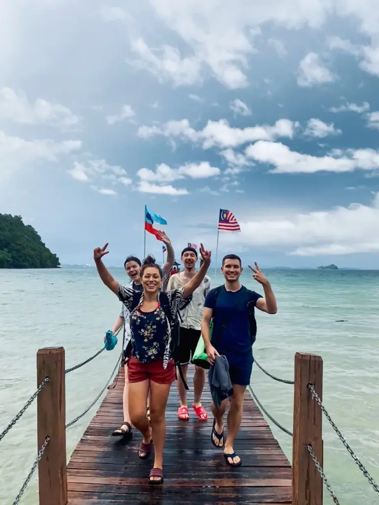Clara und Jonathan lächeln mit Freunden auf einem Holzpier, umgeben von Flaggen unter dem bewölkten Himmel Malaysias in Kota Kinabalu.