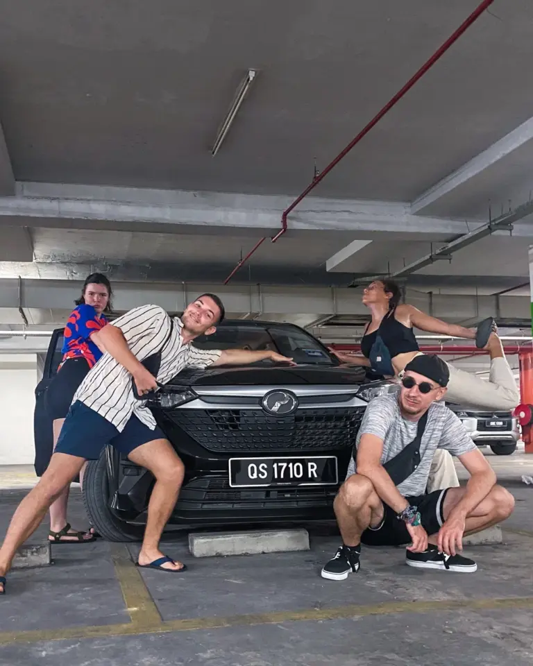 Clara und Jonathan posieren mit Freunden um ein schwarzes Auto in einem Parkhaus während eines Malaysia Roadtrips.