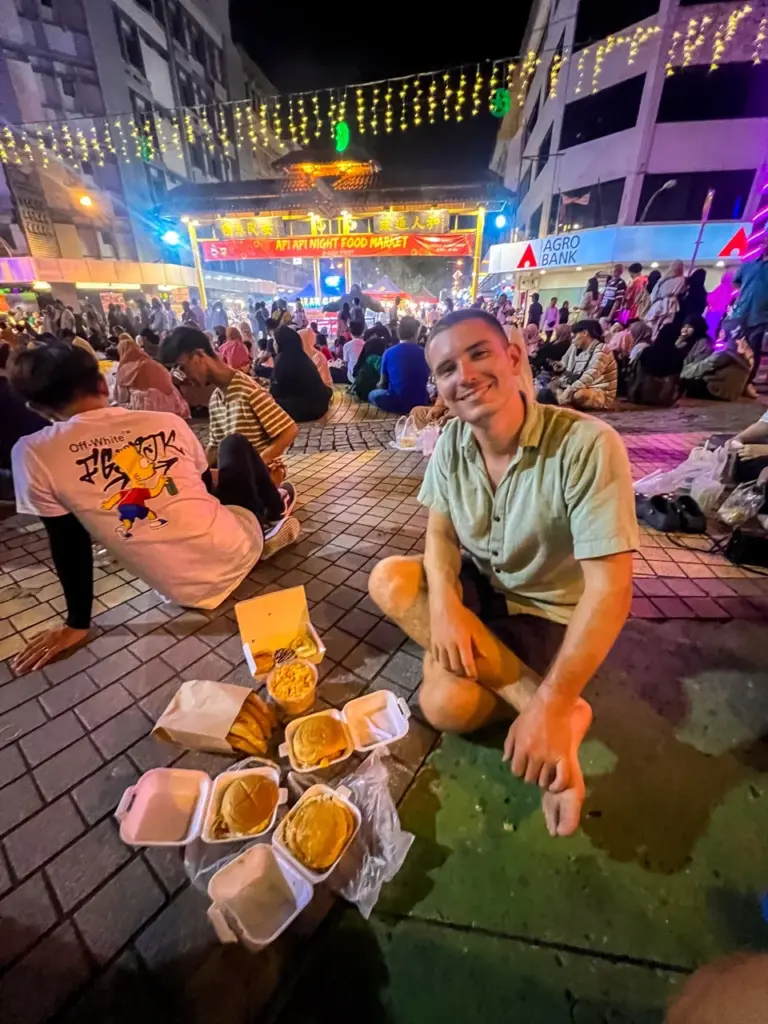 Jonathan isst Streetfood auf dem beliebten Nachtmarkt auf der Malaysia Route, umgeben von bunten Lichtern und Menschenmassen.