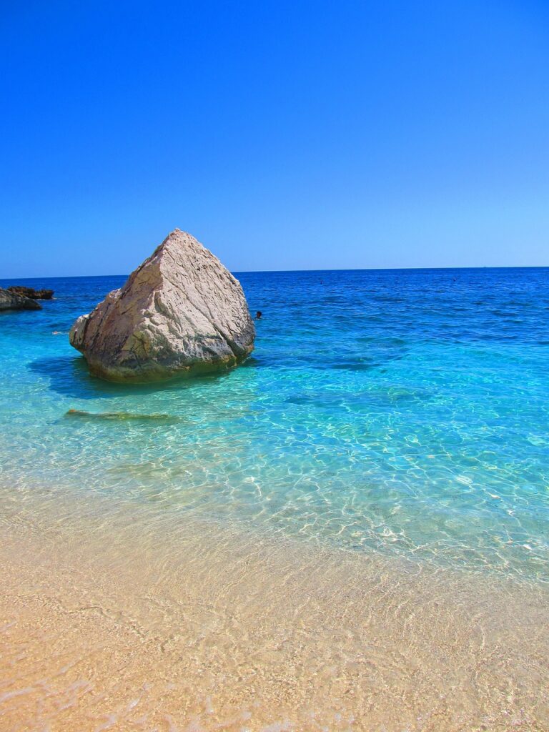 Kristallklares Wasser küsst einen Sandstrand mit einem majestätischen Felsen unter dem strahlend blauen Himmel für Ihren Sommerurlaub wohin auch immer.