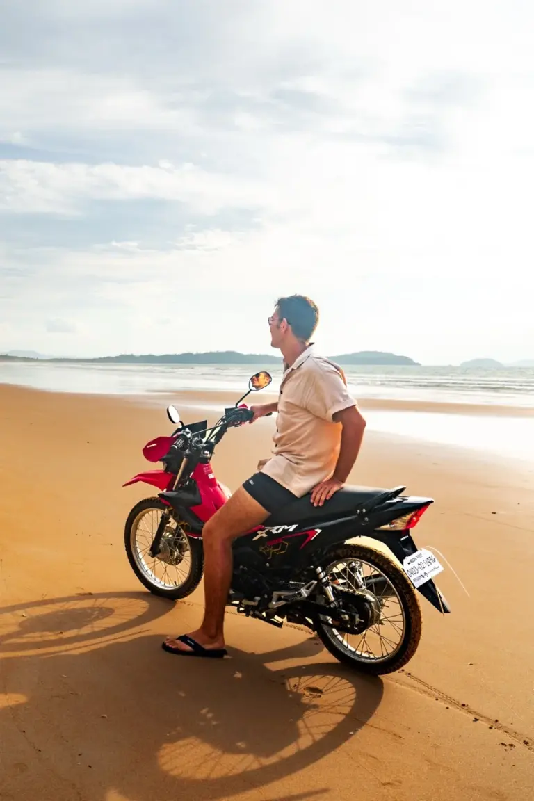 Jonathan bewundert auf einem Motorrad einen sonnigen Strand mit azurblauem Meer und träumt von seinem Reiseziel 2025.