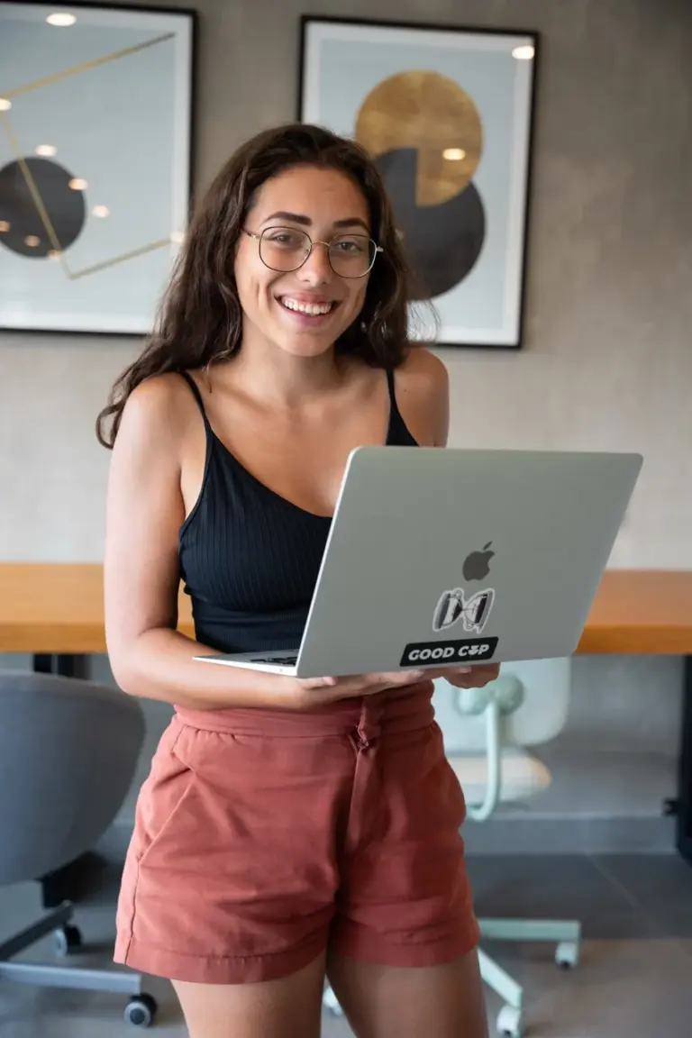 Clara, lächelnd und mit Brille, hält einen Laptop in einem kunstvoll eingerichteten Büro und plant ihre Packliste für die Weltreise.
