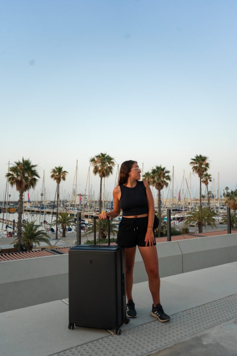 Clara in Schwarz mit Level 8 Koffer steht zwischen Palmen und Booten, bereit für ein Reiseabenteuer.