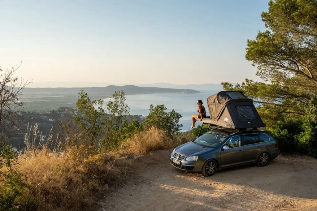 Clara sitzt im Roof Space 2 Dachzelt mit Blick auf den Sonnenuntergang, sanfte Hügel, Bäume und einen schimmernden See.