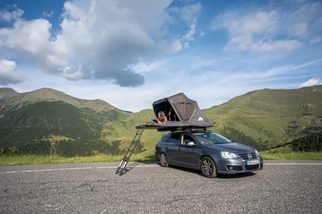 Clara sitzt im Dachzelt beim Roof Space 2 Test auf einer Bergstraße steht, umgeben von blauem Himmel im Hybrid Dachzelt.