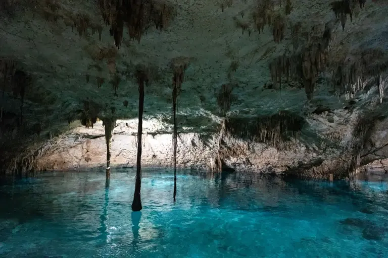 Stalaktiten und Stalagmiten hängen über dem klaren blauen Wasser in einer sonnenbeschienenen Höhle in Yucatan, Felsen ringsum.