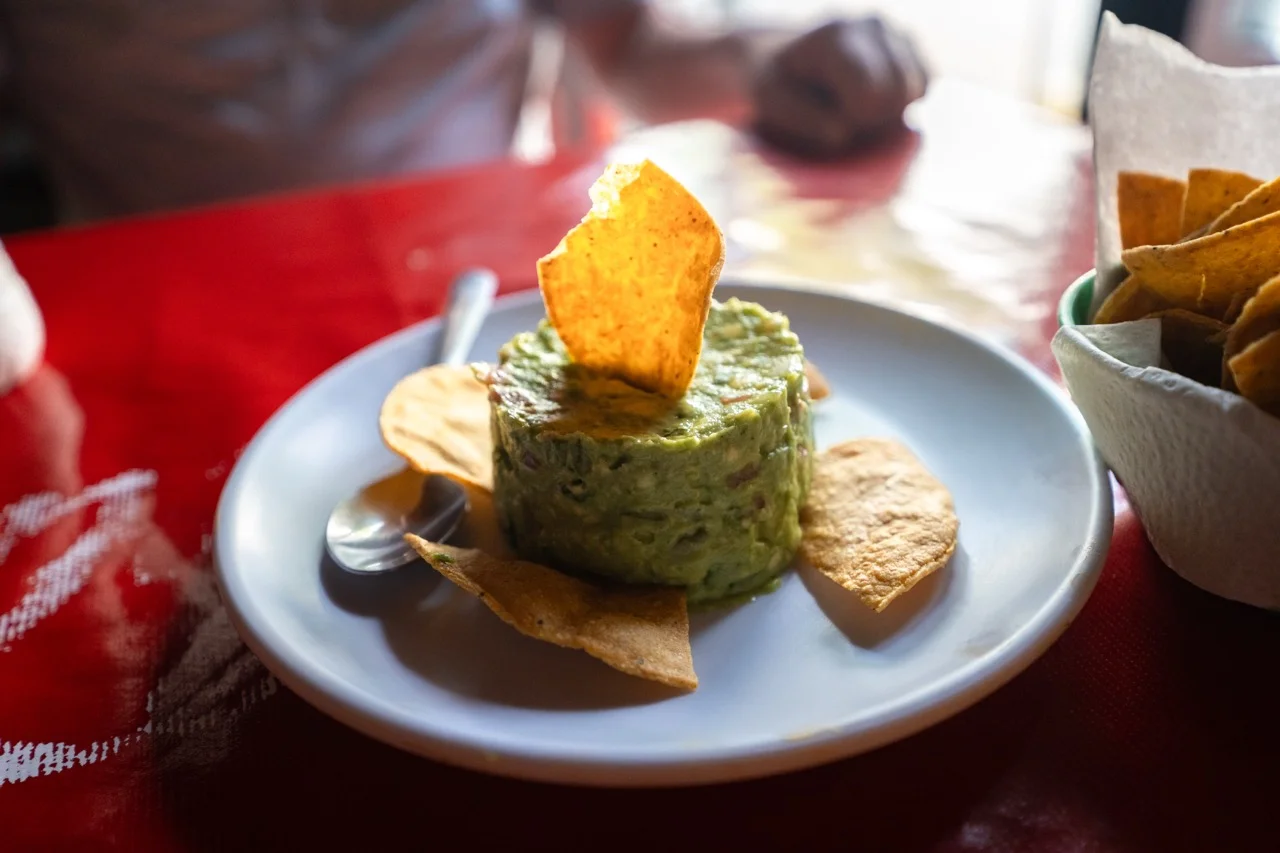 Yucatan-Guacamole mit einem Chip oben drauf, Tortilla-Chips drum herum und einem Löffel an der Seite.