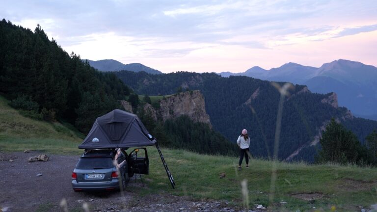 Clara steht an einem Auto mit einem Hybrid-Dachzelt und blickt auf grüne Berge bei Sonnenuntergang.