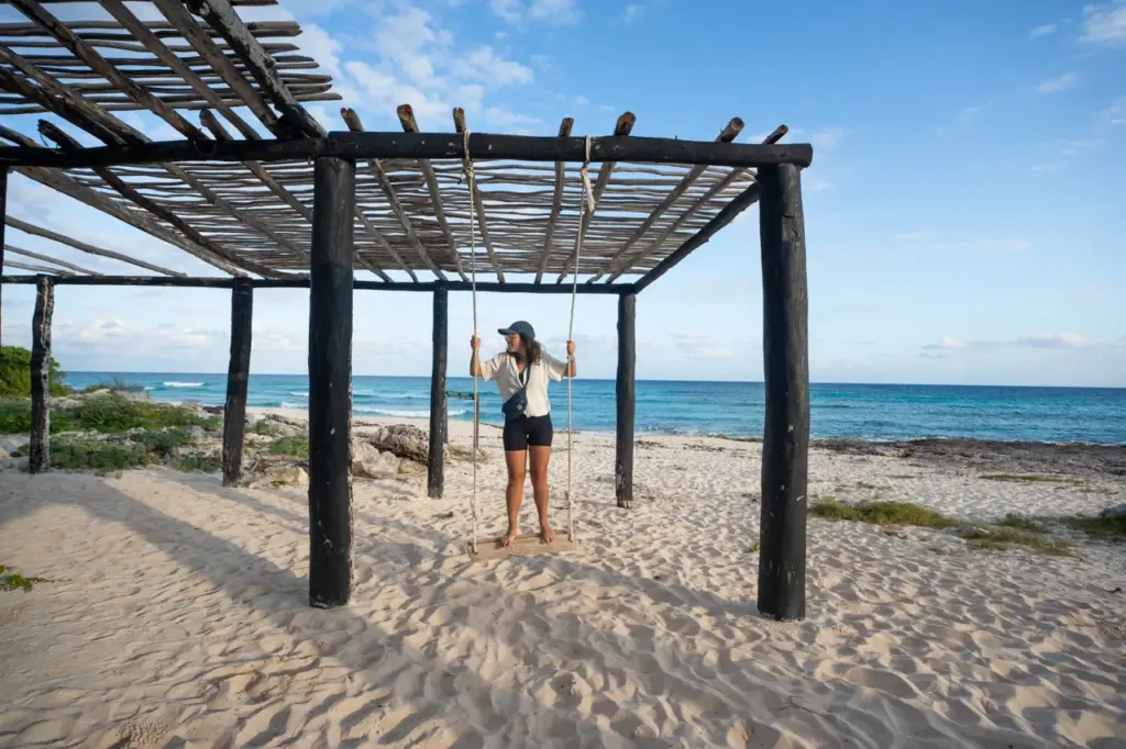 Clara in Shorts und mit Hut auf einer Schaukel unter einer Holzpergola am Strand von Playa del Carmen in Yucatan.