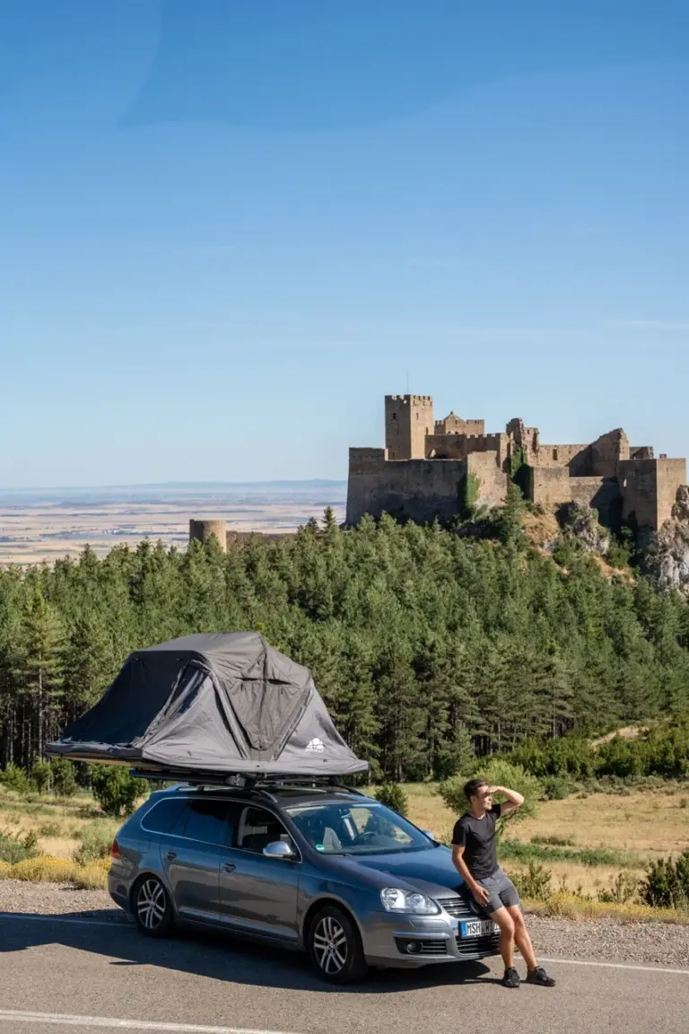 Jonathan steht bei einem Auto mit einem Roof Space Dachzelt in der Nähe einer Burg und üppigem grünen Waldes.