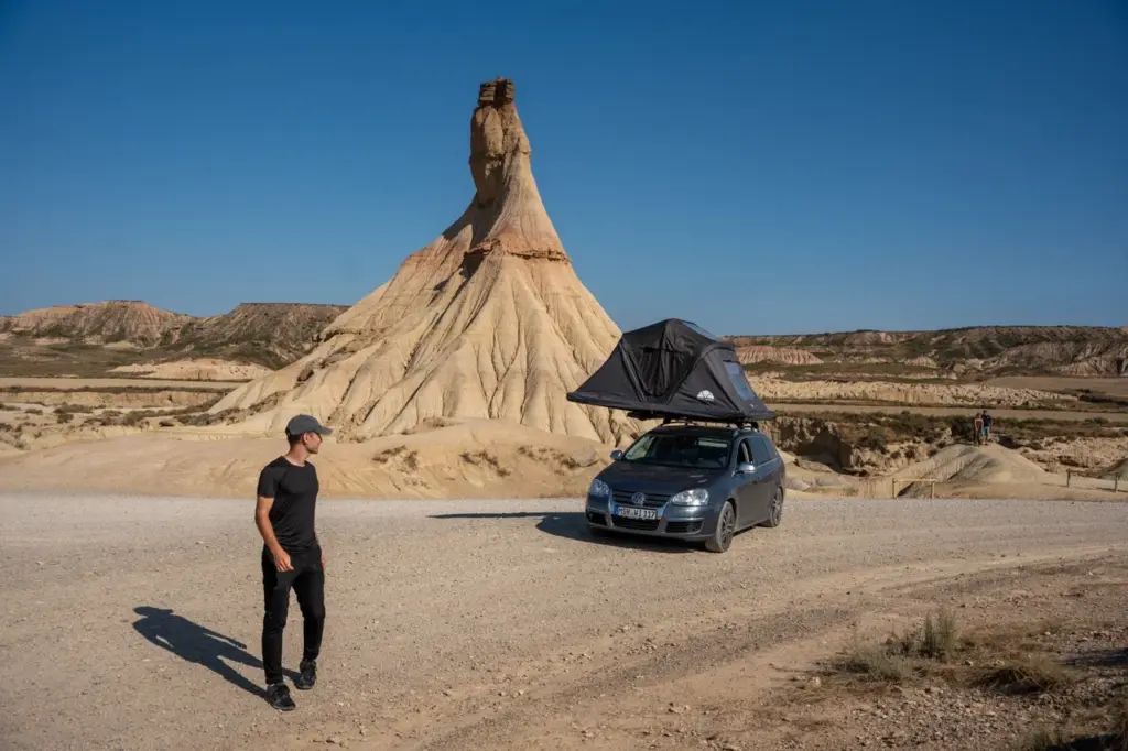 Jonathan steht neben einem Auto mit einem Roof Space Dachzelt an atemberaubenden Wüstenfelsen unter einem hellen, klaren Himmel.