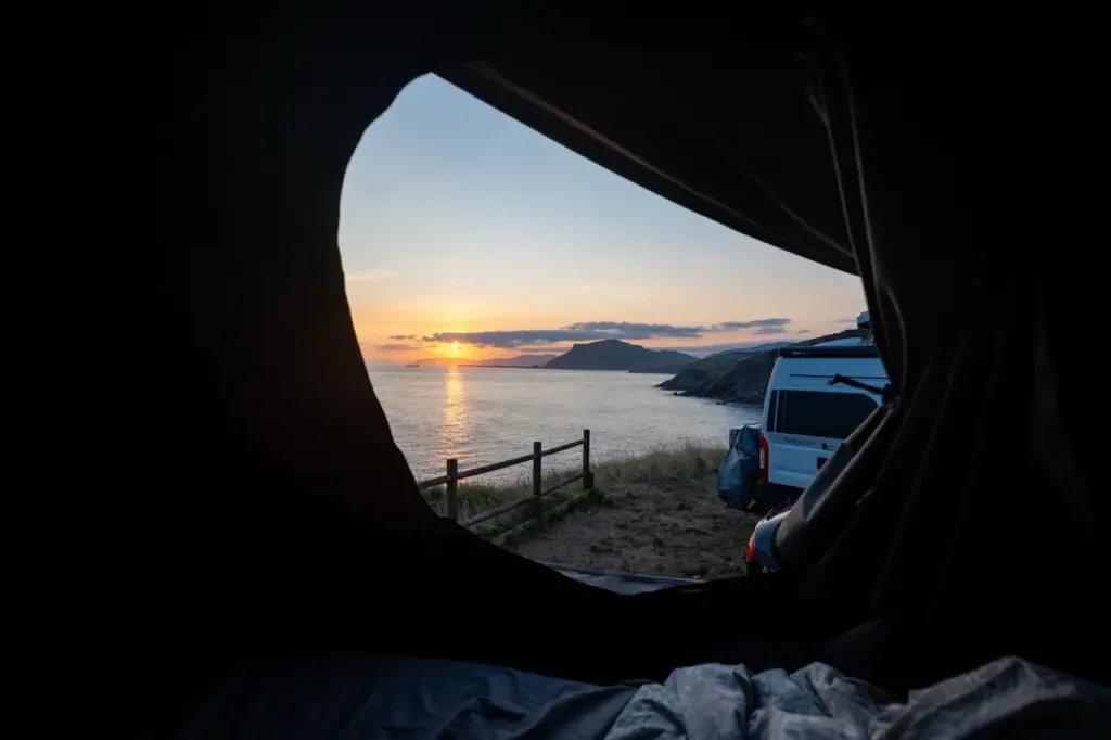 Blick auf den Sonnenaufgang aus einem Zelt an der Küste mit einem Dachzelt-Wohnmobil, das in der Nähe der Wasserkante geparkt ist.