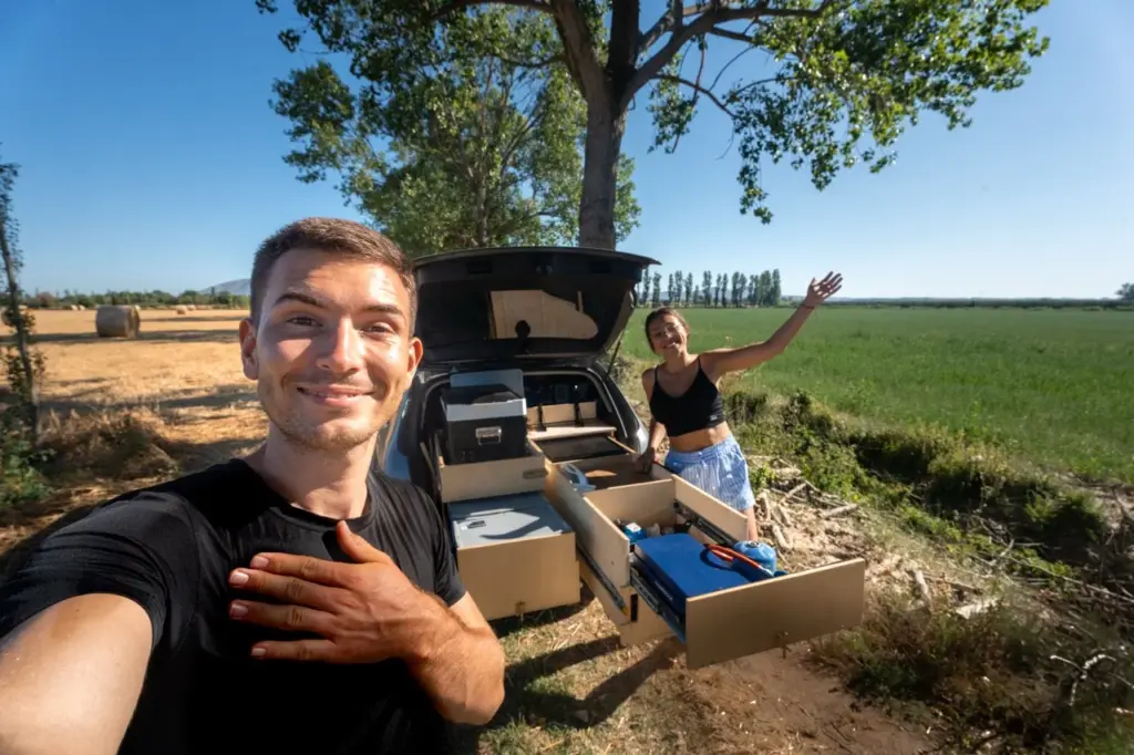 Clara und Jonathan lächeln bei ihrem Auto mit offenem Kofferraum und Dachzelt auf einer grünen Wiese.