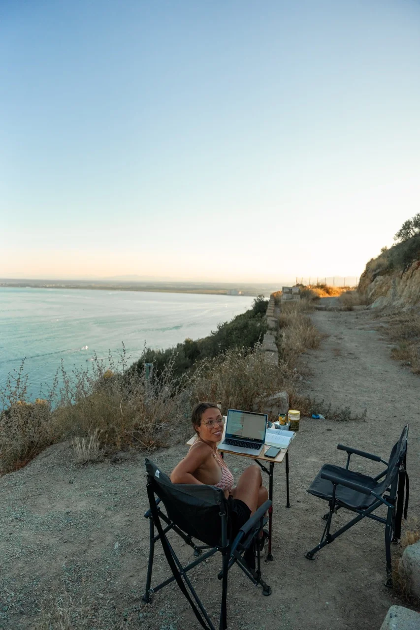 Clara sitzt bei Sonnenuntergang am Meer neben dem Dachzelt auf einem Klippenweg