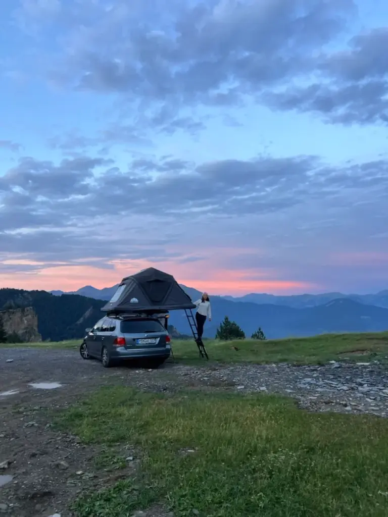 Clara steht bei Sonnenuntergang neben einem Auto mit einem Dachzelt und blickt auf grasbewachsene Hügel und Berge.