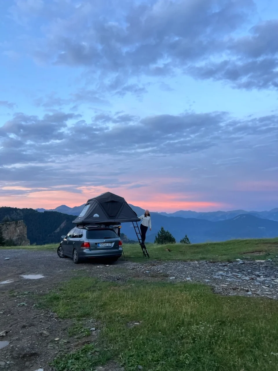 Clara steht bei Sonnenuntergang neben dem Auto mit Dachzelt und blickt auf grüne Hügel und Berge