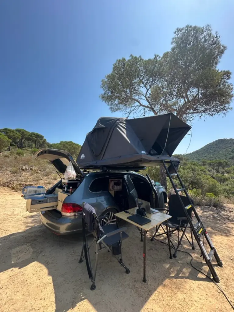 Auto mit Dachzelt, Campingausrüstung und Stühlen, die an einem sonnigen, grünen Platz in der Natur aufgestellt sind.