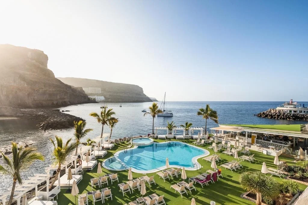 Der Pool des Resorts ist von Palmen gesäumt und bietet einen Blick auf die ruhige Bucht und die Klippen von Gran Canaria unter blauem Himmel.