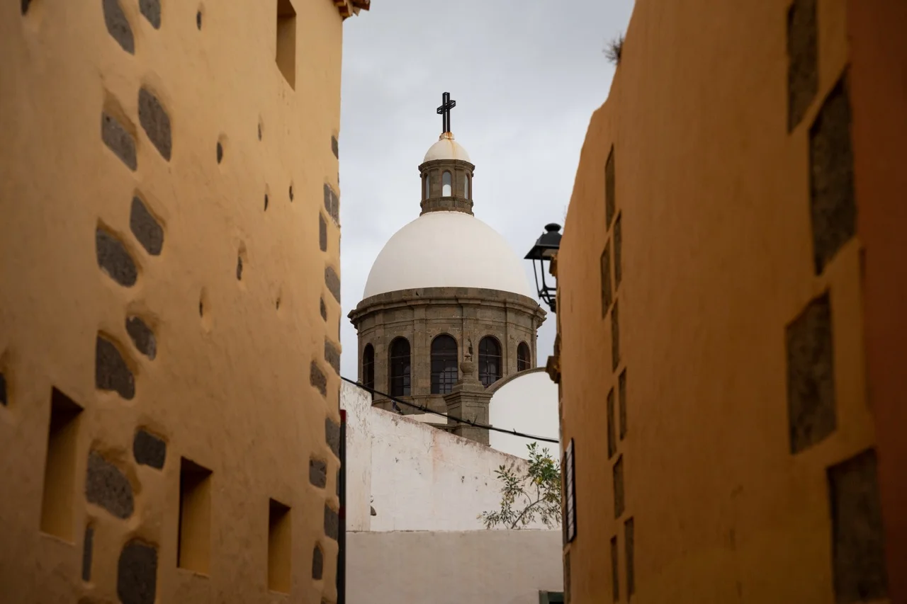 Die Kuppel der Kirche San Juan Bautista erhebt sich auf Gran Canaria, umrahmt von gelb-orangenen Gebäuden und Wolken.