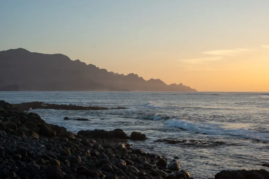 Die felsige Küste von Gran Canaria bei Sonnenuntergang, ruhige Wellen, ferne Berge und ein klarer, leuchtender Himmel.
