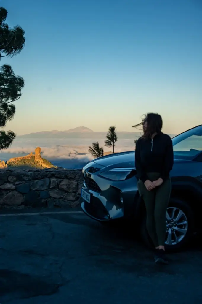 Clara an ihrem Auto auf einer Bergstraße auf Gran Canaria bei Sonnenuntergang, Wolken und felsige Gipfel im Hintergrund.
