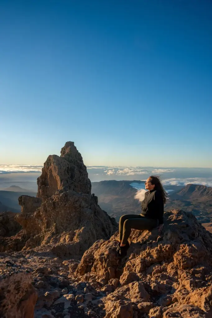 Clara sitzt auf felsigem Grund in Gran Canaria, mit den Bergen von Maspalomas und den Wolken des Sonnenuntergangs in der Ferne.