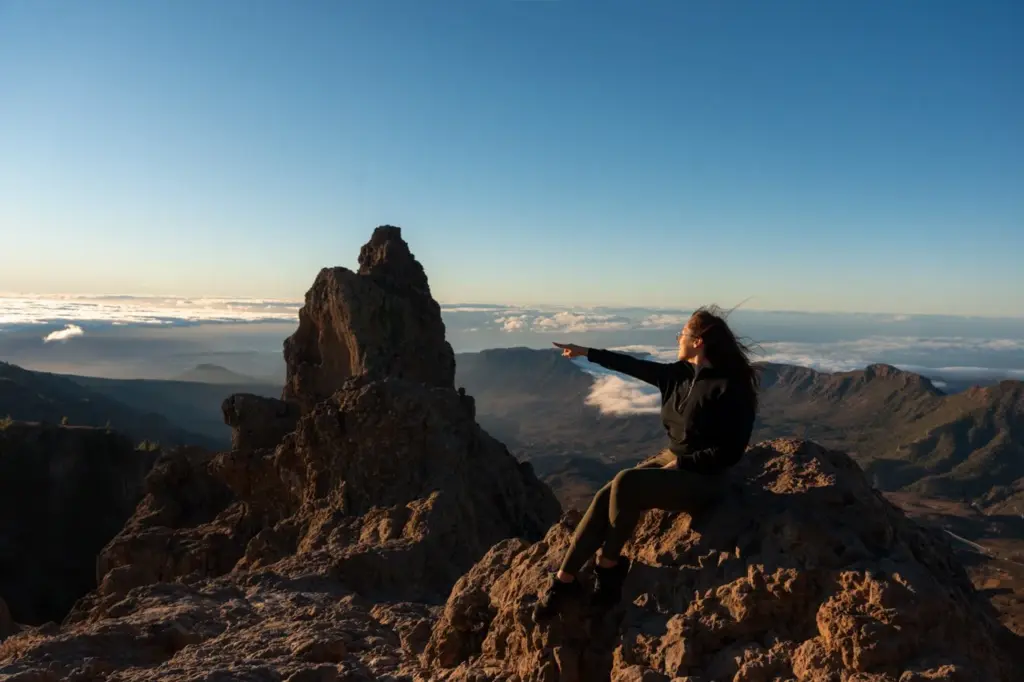 Clara sitzt auf einem felsigen Gipfel auf Gran Canaria. Der Roque Nublo ist unter einem strahlend blauen Himmel zu sehen.