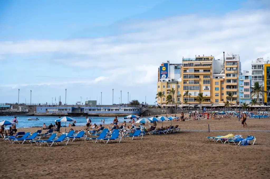 Blaue Liegestühle säumen den Sandstrand von Gran Canaria, mit bunten Häusern und wolkenverhangenem Himmel in der Nähe.