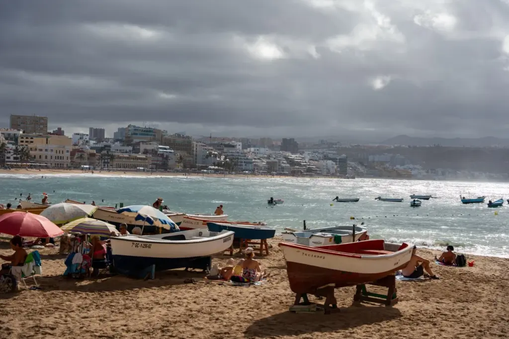Bunte Boote am Sandstrand von Las Canteras auf Gran Canaria, dahinter Gebäude der Stadt und bewölkter Himmel.