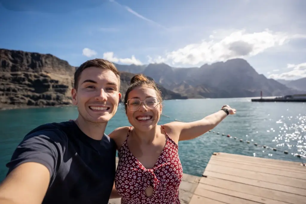 Clara und Jonathan lächeln an einer Bucht, während die Berge Gran Canarias unter dem sonnigen Himmel leuchten.