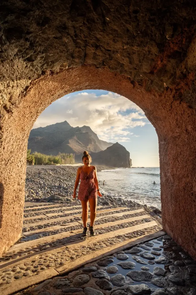 Clara in einem roten Kleid steht bei Sonnenuntergang unter einem steinernen Torbogen am Strand von Gran Canaria.